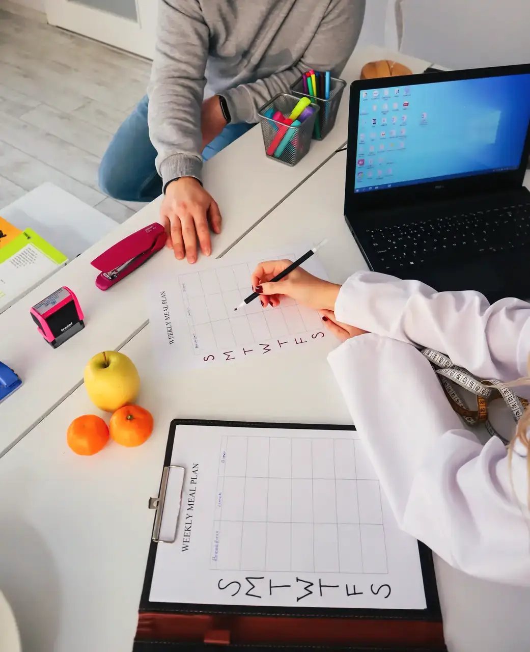 A dietitian and client working through a weekly meal chart at a desk with fresh fruit